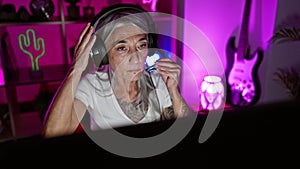 Serious yet beautiful, middle-aged, grey-haired woman streamer wearing headphones, stretching arms in dark gaming room amidst