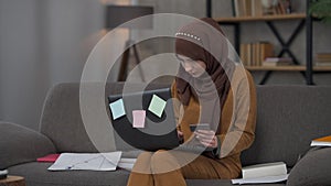 Serious beautiful Middle Eastern woman in hijab holding business card typing on laptop keyboard. Portrait of young