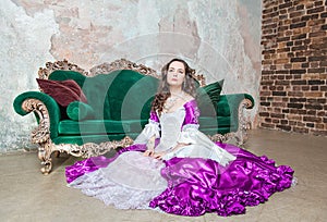 Serious beautiful woman in fantasy white and purple rococo style medieval dress sitting on the floor near sofa