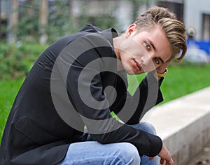 Serious blond young man in jeans and jacket, sitting outdoors