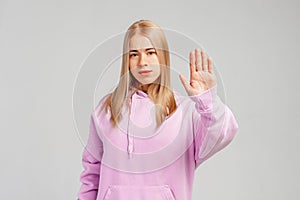 Serious blonde girl showing stop sign and frowning displeased, telling no, refusing something, standing in trendy hoody over gray