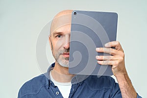 Serious bold man cover half of his face with digital tablet looking at camera isolated on white background. Posting bad