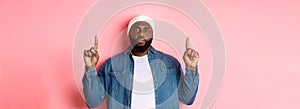 Serious and bothered Black man with beard, staring at camera and pointing fingers up, showing promo, standing over pink