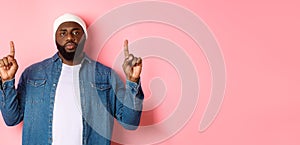 Serious and bothered Black man with beard, staring at camera and pointing fingers up, showing promo, standing over pink