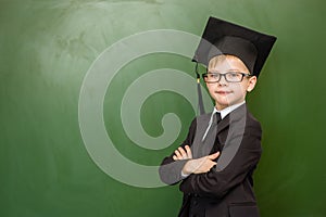 Serious boy in graduation cap standing near green chalkboard