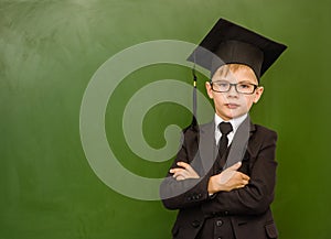 Serious boy in graduation cap standing near green chalkboard