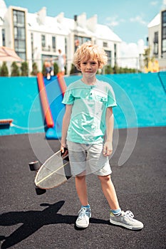 Serious boy holding his skateboard in front of sports ground.