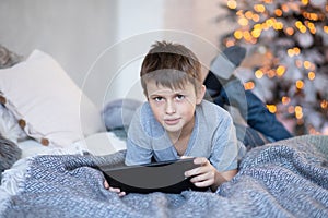 A serious boy is holding a tablet in his hands. The child plays computer games on the tablet. The boy lies on the bed opposite the