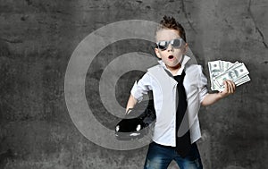 Serious boy little businessman in white shirt, jeans, tie and sunglasses standing and holding toy car present and dollars cash