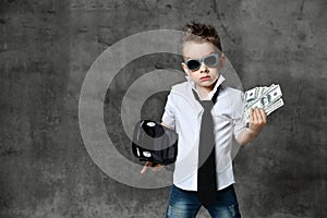 Serious boy little businessman in white shirt, jeans, tie and sunglasses standing and holding toy car present and dollars cash