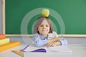 Serious boy sitting at desk with apple on his head in classroom. Adorable child going back to school
