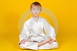 Serious boy in a white kimono sitting on a yellow background. Martial arts. Sports since childhood. Little boy sitting in lotus