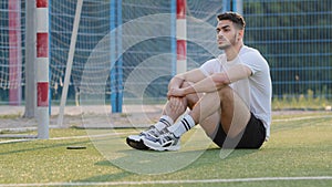 Serious, brooding Middle Eastern footballer sitting on grass of soccer field against goal, resting after game or