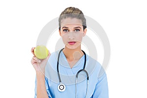 Serious brown haired nurse in blue scrubs holding a green apple