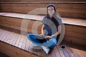 A serious brunette student girl , sits on wooden steps, with an open laptop, typing on a keyboard, learns online.