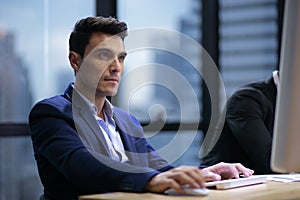 Serious business man in suit sitting at desk using computer in office on window. tired worker working overtime in evening .