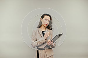 Serious business woman in a jacket stands with a paper tablet and a pen in his hands and looks at the camera on a beige background