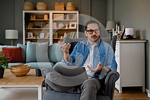 Serious businessman gesturing at camera while working over laptop on armchair in home office