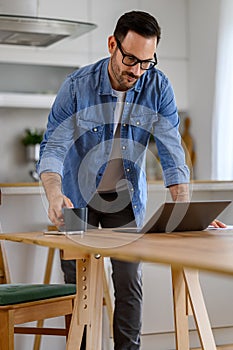 Serious businessman holding coffee cup and working over laptop while standing at desk in home office