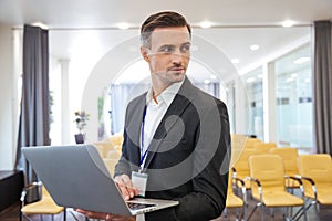 Serious businessman holding laptop in empty meeting hall