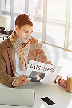 Serious businessman sitting at table, holding paper cup and reading business newspaper