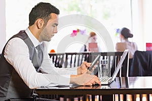 Serious businessman using phone while working on laptop