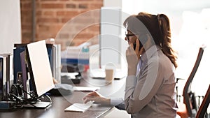 Serious businesswoman in glasses using computer, talking on phone