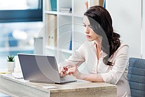 Serious businesswoman sitting at desk and working with laptop in office