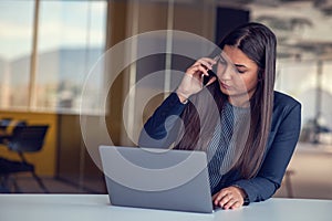 Serious businesswoman talking on mobile phone at office workplace, working at computer. Secretary answering telephone