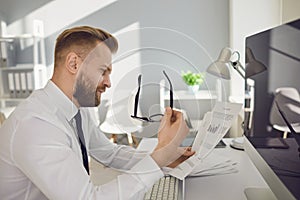 Serious busy businessman reads working documents sitting at a table with a computer in the office.