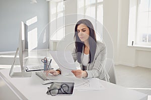 Serious busy businesswoman reads working documents sitting at a table with a computer