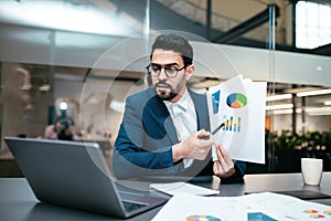 Serious busy millennial arab guy with beard in glasses, suit sits at table shows color chart in computer webcam