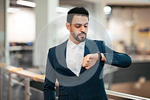 Serious calm attractive young arab ceo man with beard in suit looks at smart watch wait for client in office interior