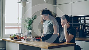 serious caucasian couple in kitchen, woman is cutting salad, she is standing silent, man is sitting at table working on