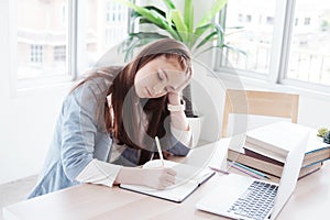 Serious Caucasian girl student is writing on notebook. She so bored about online learning and doing many homework with laptop at