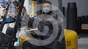 Serious Caucasian man in coronavirus face mask standing at forklift in warehouse looking at camera. Portrait of