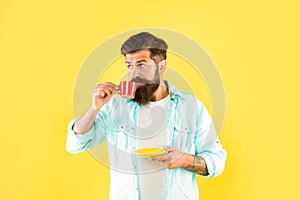 Serious caucasian man drinking coffee from cup holding saucer yellow background, caffeine