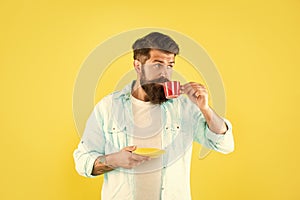 Serious caucasian man drinking coffee from cup holding saucer yellow background, caffeine