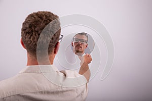 Serious caucasian man looks at himself in the mirror and reflects on life. Pensive person with eyeglasses. Guy isolated