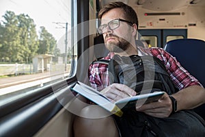 Serious Caucasian man rides a train and looks thoughtfully out the window, in hands he holds a book and a pencil.