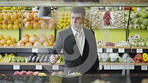 Serious Caucasian man in suit choosing kiwi fruit in grocery. Portrait of confident adult businessman buying berry in