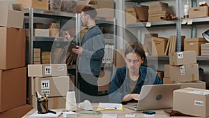 Serious caucasian man typing parcels details at the keyboard at the laptop while his male caucasian colleague sorting