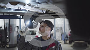 Serious Caucasian man in workwear and cap walking under car bottom with flashlight. Male adult auto mechanic examining