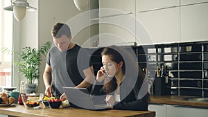 Serious caucasian nice couple in kitchen, man is cutting salad, he is standing silent, woman is sitting at table working