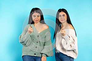 Serious caucasian twin girls show their fist and look at the camera. Blue background.