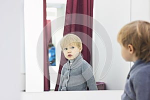 Serious Child - European blond boy tries on clothes in a store in the dressing room and looks at himself in the mirror