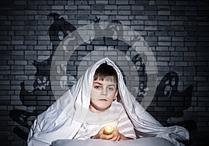 Serious child with flashlight hiding under blanket