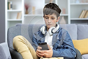 Serious child of a teenage boy in headphones sitting at home on the sofa, using the phone. Listens to music, watches