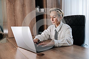 Serious concentrated attractive middle-aged business woman sitting in modern light office at desk working on laptop