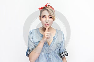 Serious concentrated blonde woman wearing blue denim shirt with finger near lips, keeps silence.